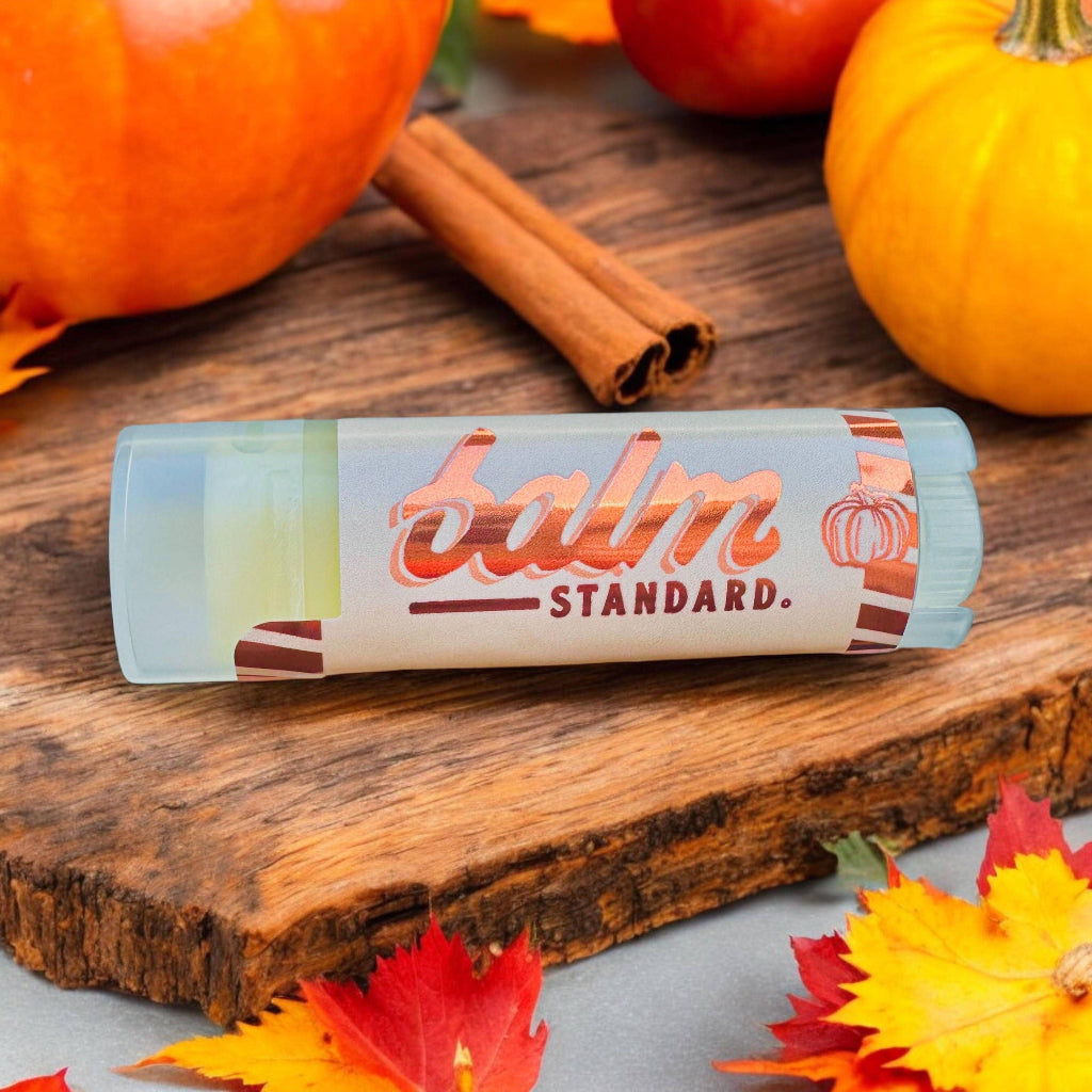 A tube of Balm Standard lip balm placed on a rustic wooden board, surrounded by autumn-themed props including pumpkins, cinnamon sticks, and fall leaves.