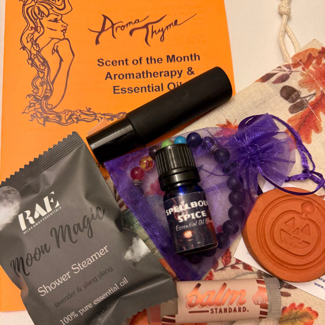 A curated autumn-themed aromatherapy and essential oil gift set arranged on a white surface. The set includes an orange Aroma Thyme 'Scent of the Month' card, a black spray bottle, a gray RAE 'Moon Magic' shower steamer packet, a small blue 'Spellbound Spice' essential oil bottle, a terracotta pumpkin diffuser, a purple chakra beaded bracelet in a sheer purple bag, and a pumpkin spice Balm Standard stick.