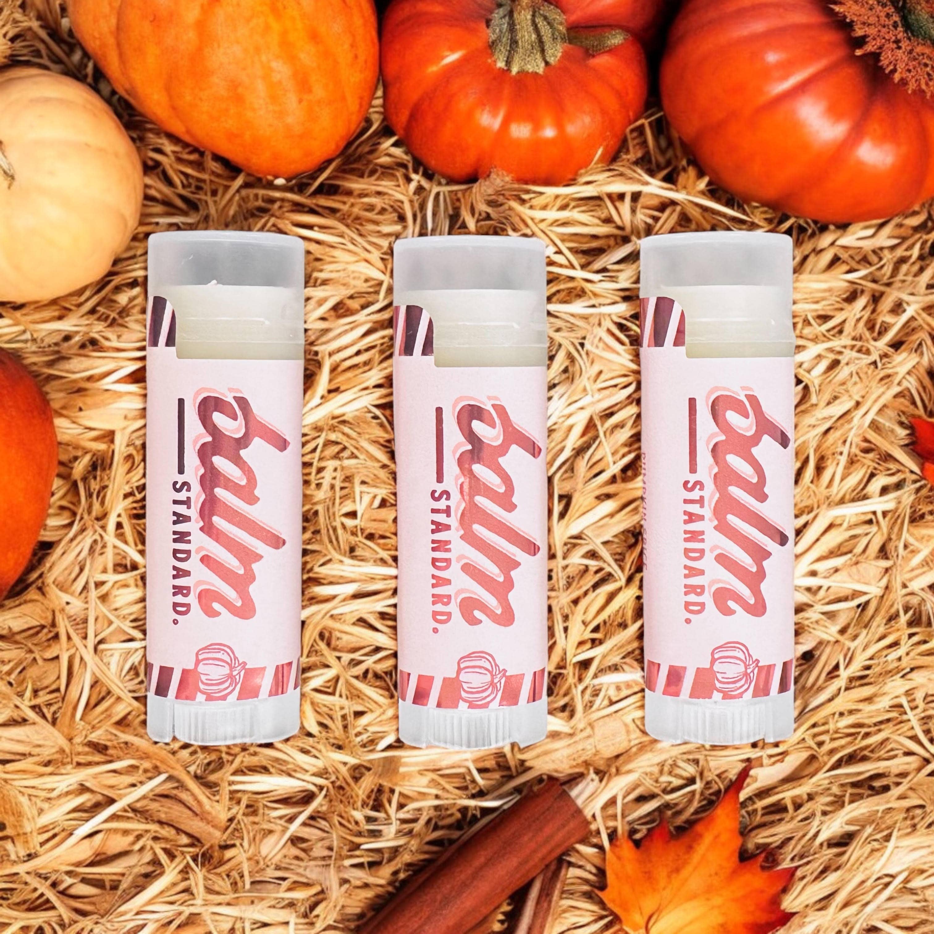 Three tubes of Balm Standard Pumpkin Spice Lip Balm arranged on a bed of straw with pumpkins, cinnamon sticks, and a fall leaf around them.